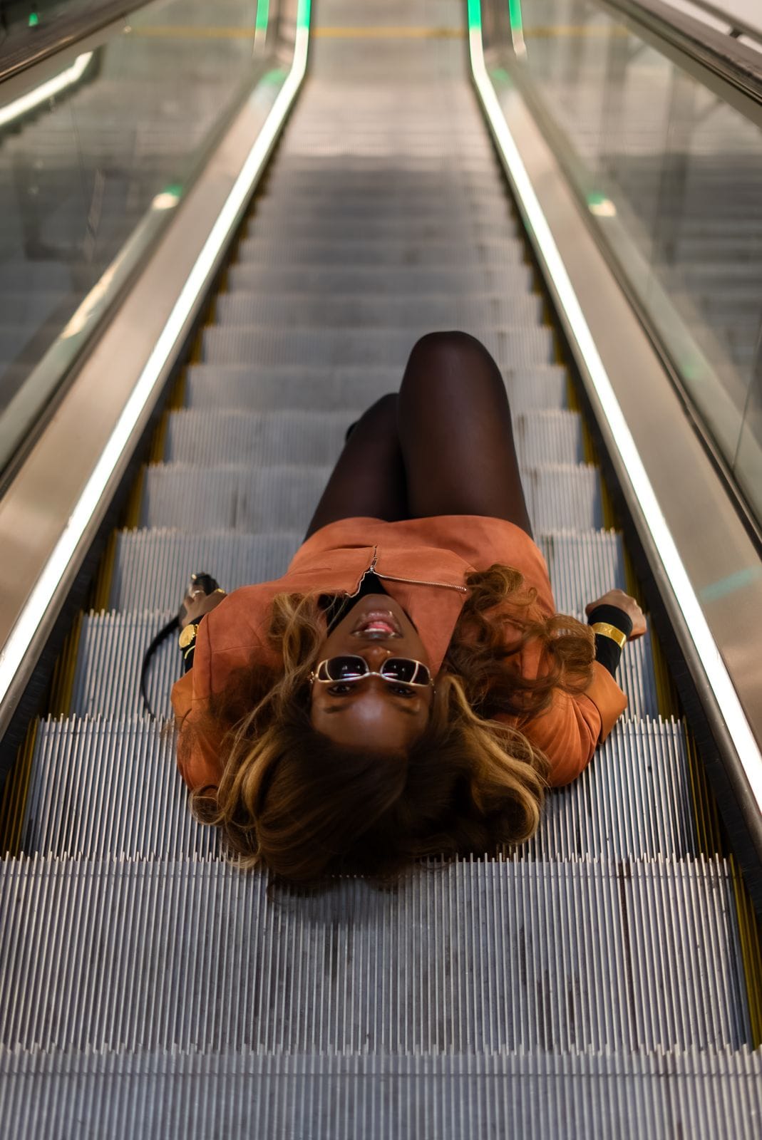 Personne allongée sur un escalator, veste orangée, lunettes de soleil.