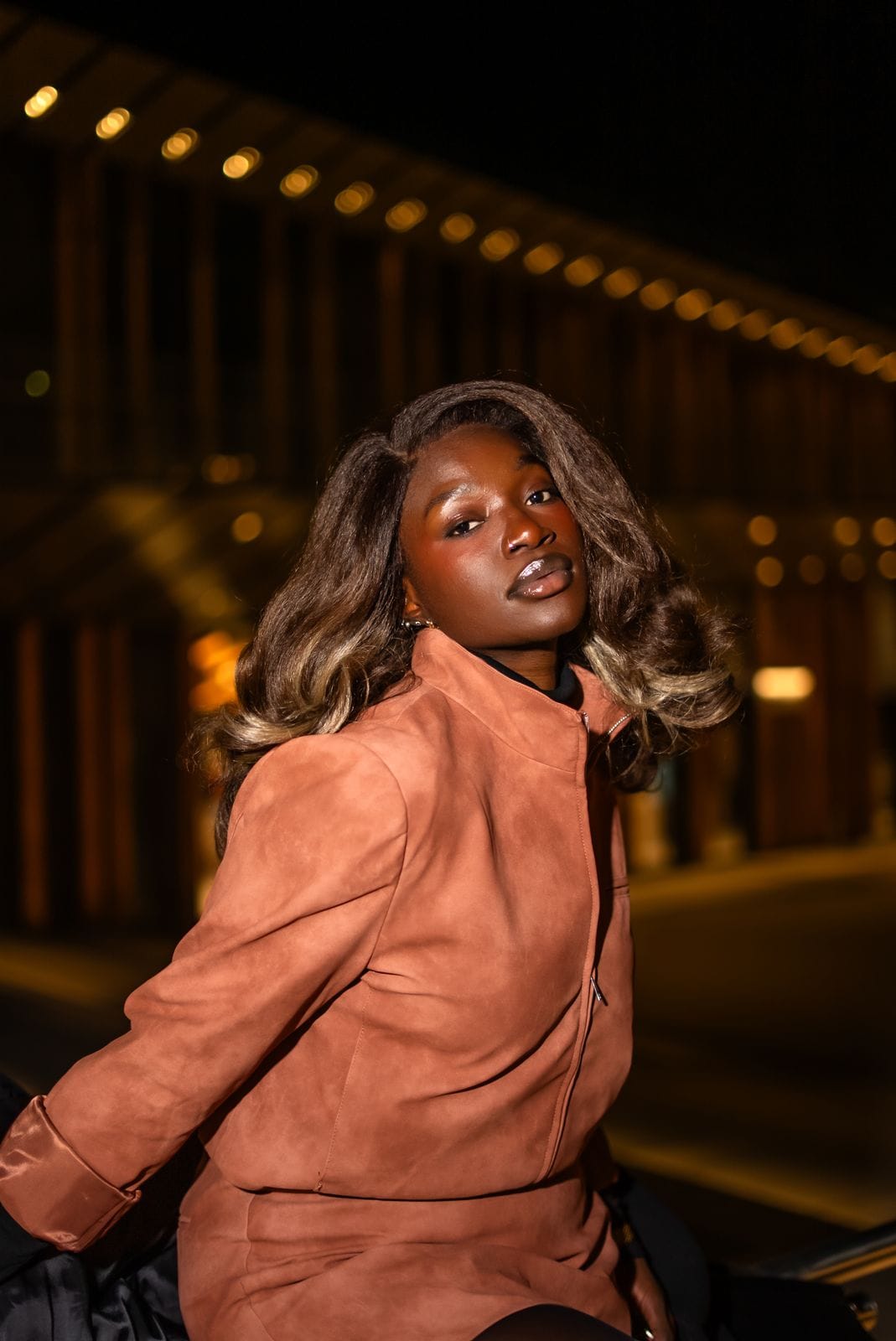 Femme en veste en daim caramel, de nuit, devant des lumières urbaines.
