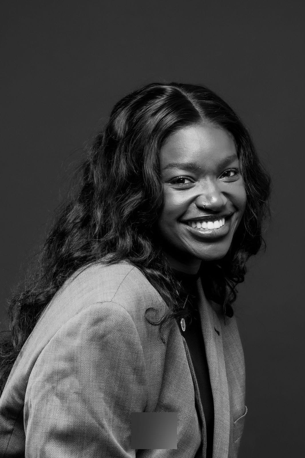 Femme souriante aux cheveux ondulés, veste grise, fond sombre, en trois quarts.