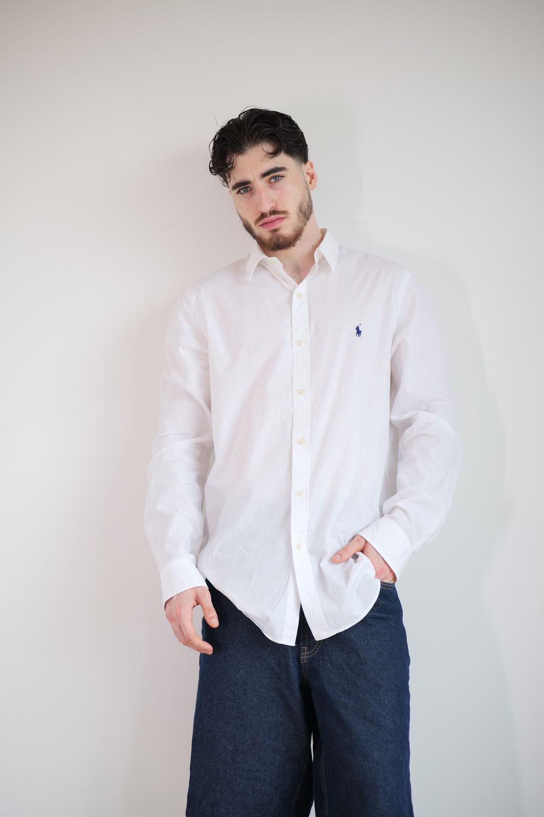 Homme en chemise blanche et jean sombre, devant un fond clair.