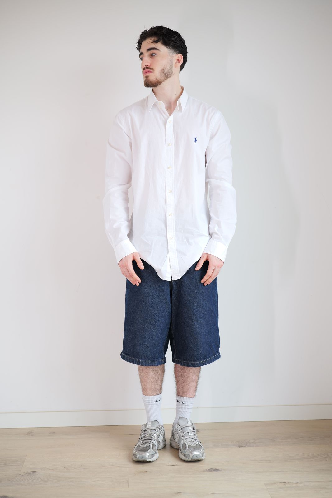 Personne en chemise blanche et bermuda denim, debout devant un mur blanc.