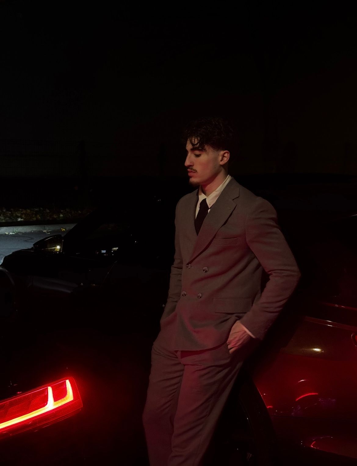 Homme en costume croisé, adossé à une voiture sombre, la nuit.