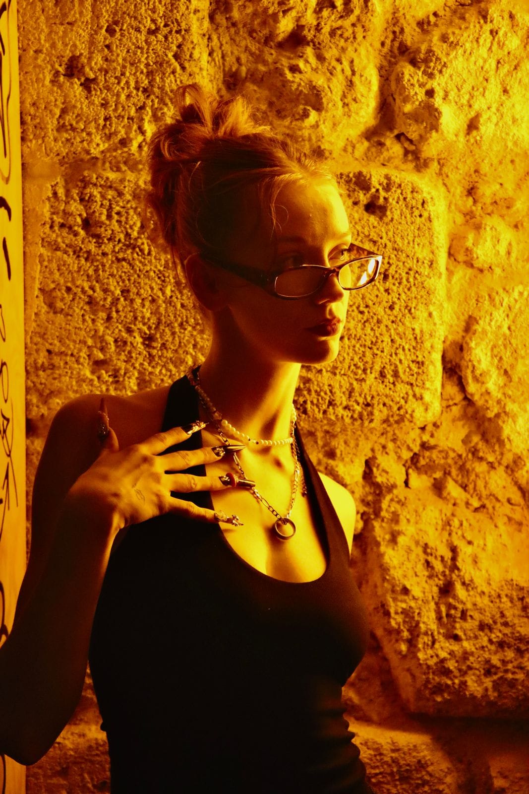 Femme aux lunettes, colliers superposés, mur de pierre éclairé en ambre.