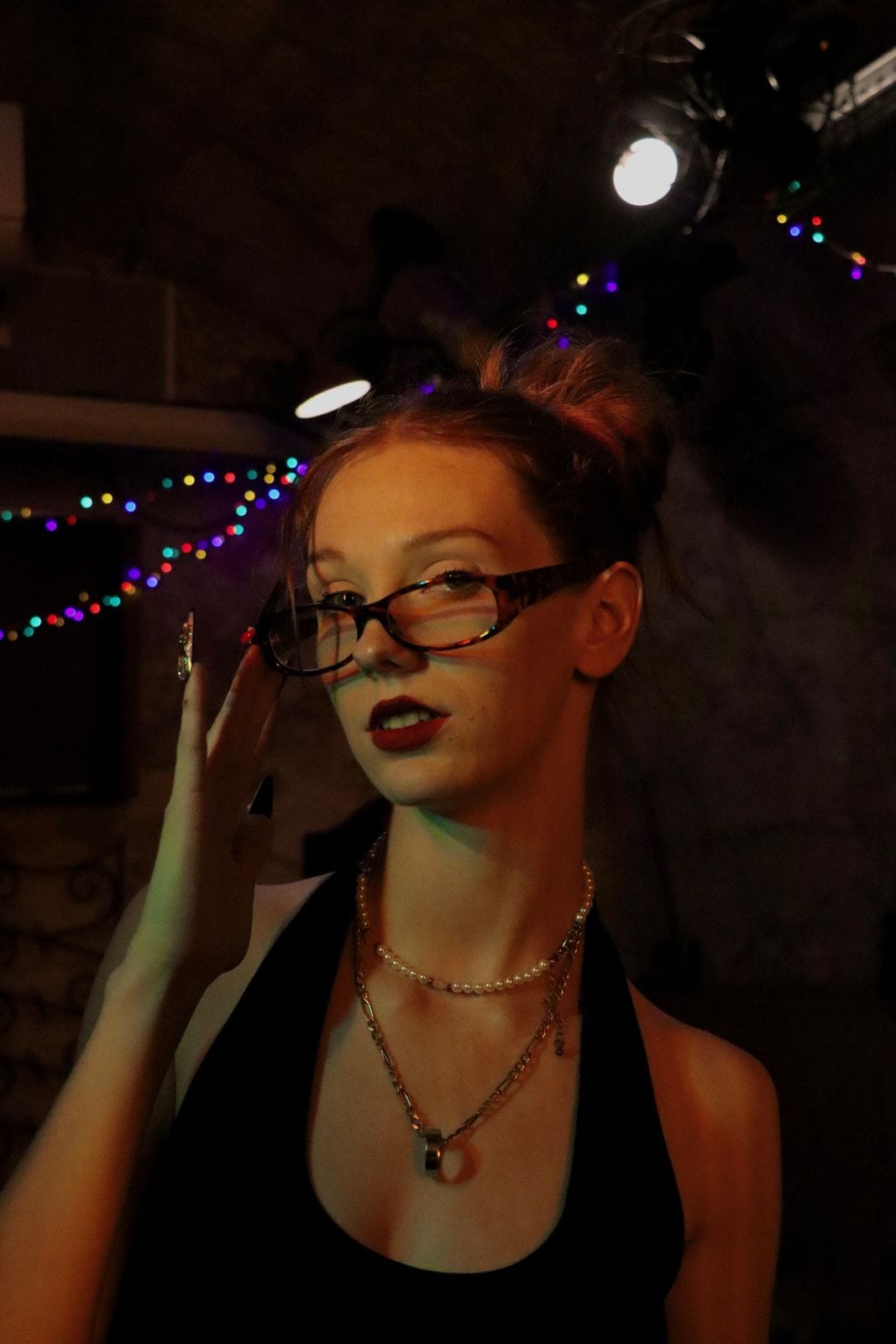 Femme à lunettes écaille, regard par-dessus la monture, lumières colorées en arrière-plan.
