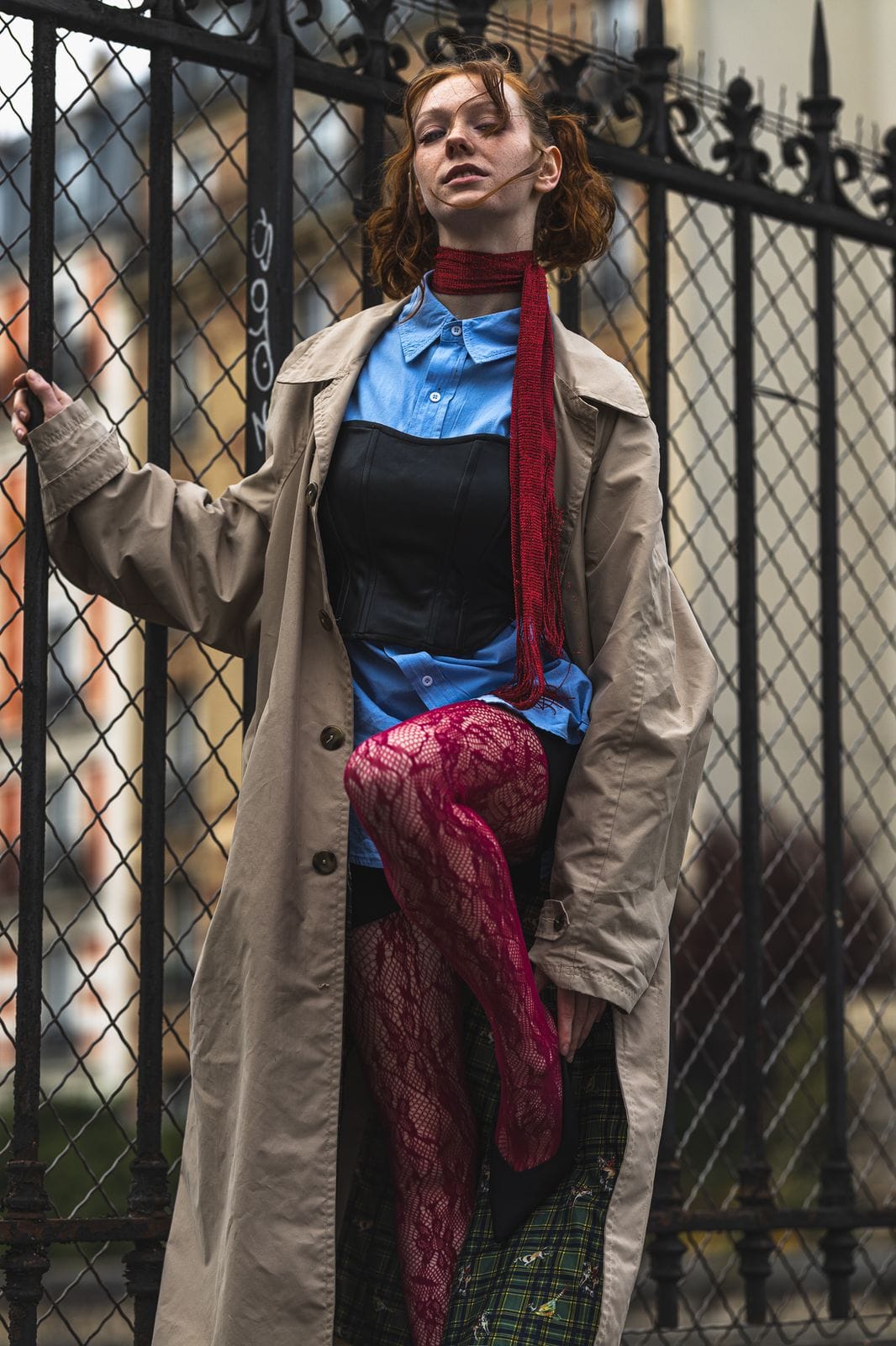 Femme rousse en trench beige, adossée à une grille noire, jambe relevée.
