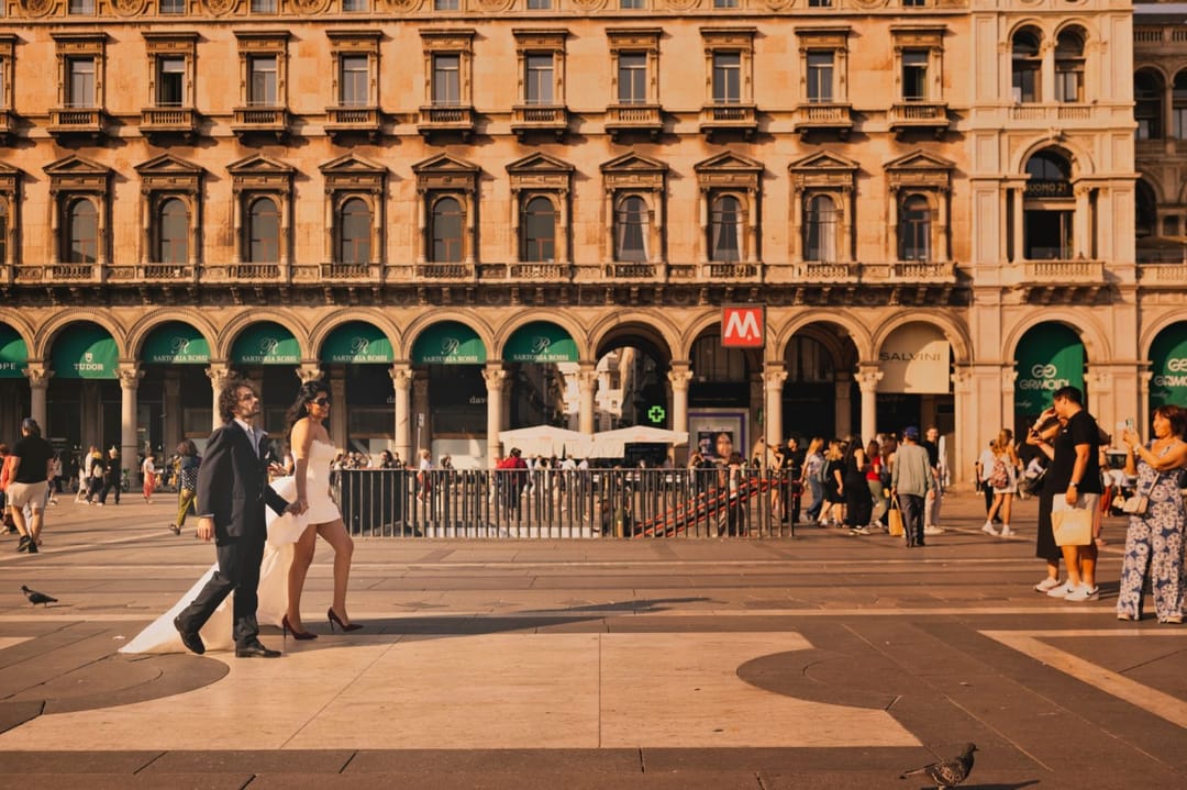 Un couple en tenue de mariage marche sur une place ensoleillée.