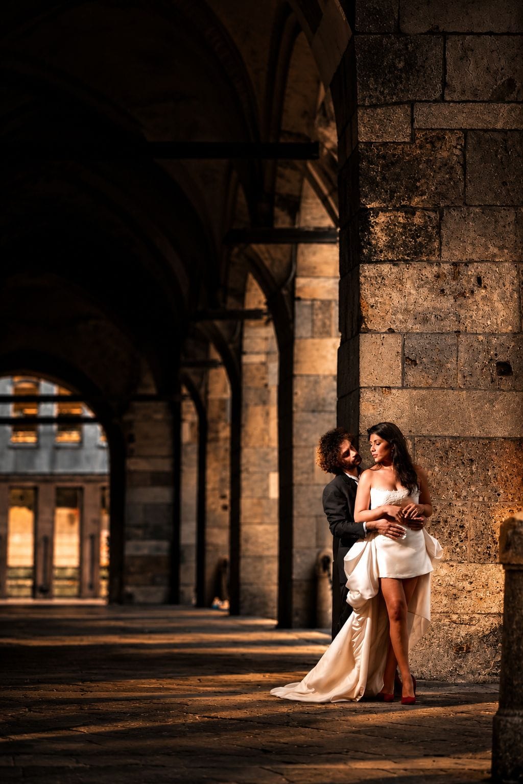 Couple enlacé sous une arcade de pierre, lumière dorée, robe blanche.