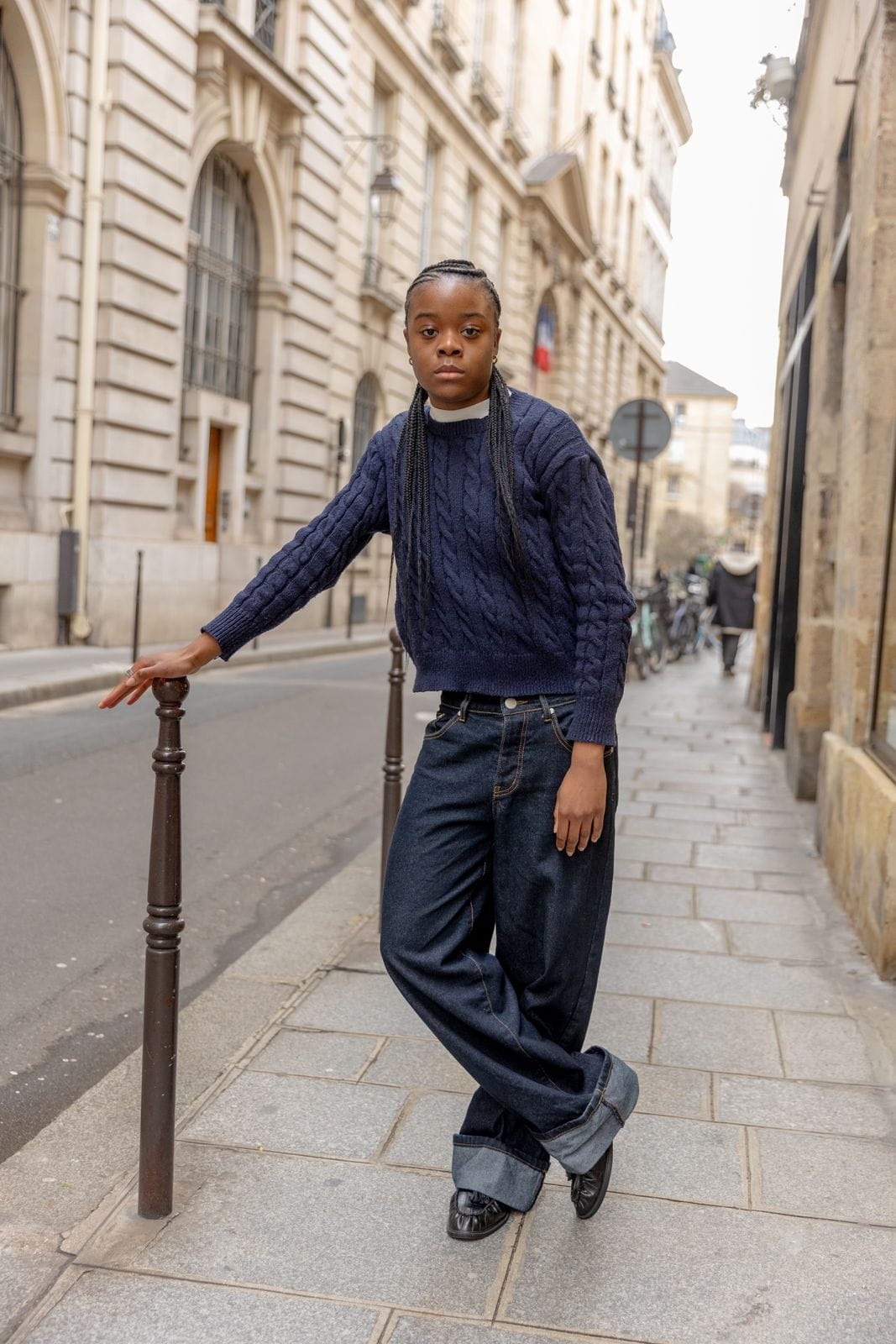 Personne debout près d'une barrière, pull torsadé bleu marine, sur un trottoir.