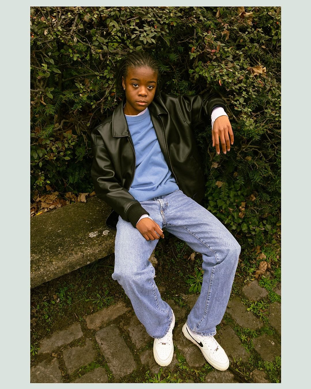 Personne assise sur un muret, devant une haie, veste en cuir.